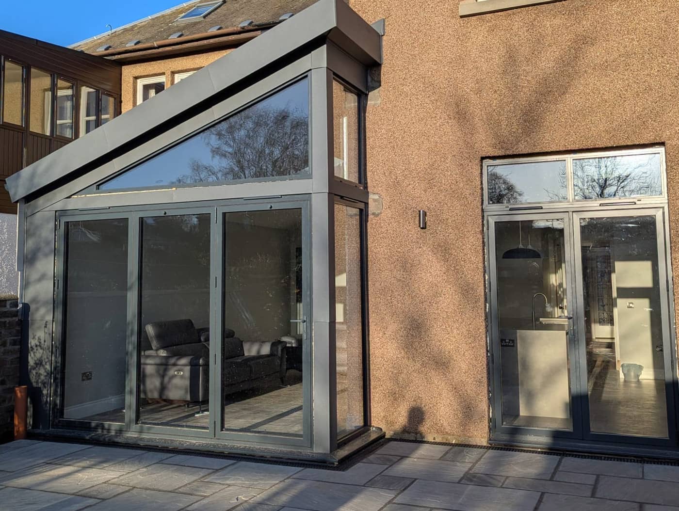 Completed single-storey rear extension with a mono-pitch roof, full-height glazed gable and grey aluminium bi-fold doors, beside new glazed doors into the house